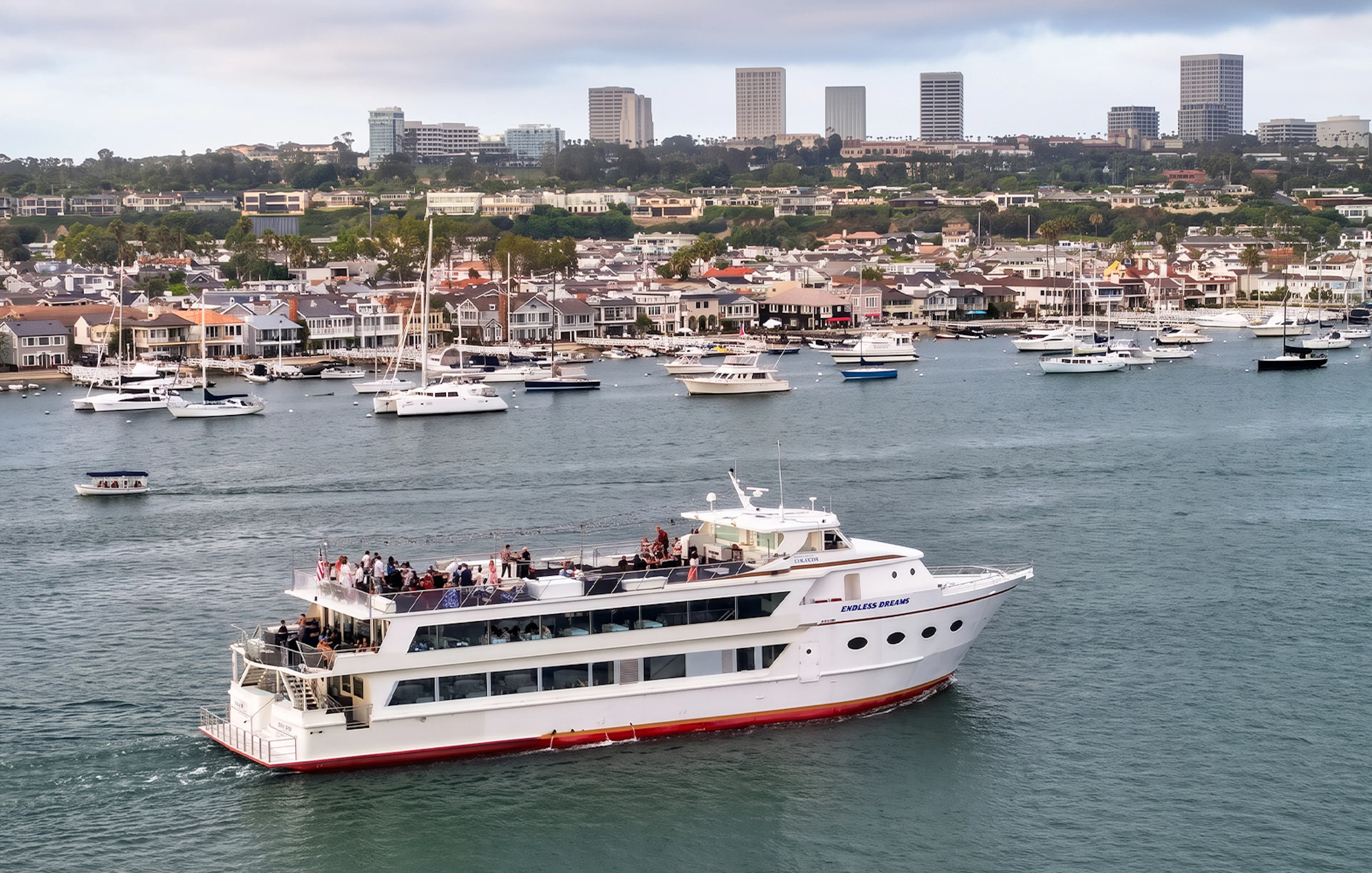 Cruise ship with guests on deck in Newport Beach harbor, city skyline in background.
