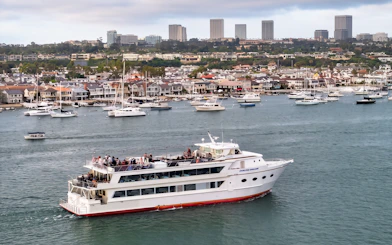 Cruise ship with guests on deck in Newport Beach harbor, city skyline in background.