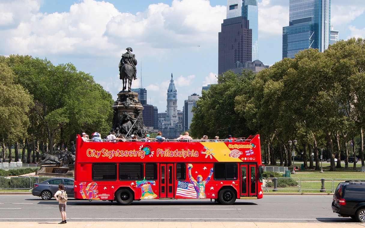 Red double-decker bus on Philadelphia Hop-on Hop-off Tour near Eakins Oval with city skyline.