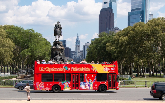 Red double-decker bus on Philadelphia Hop-on Hop-off Tour near Eakins Oval with city skyline.