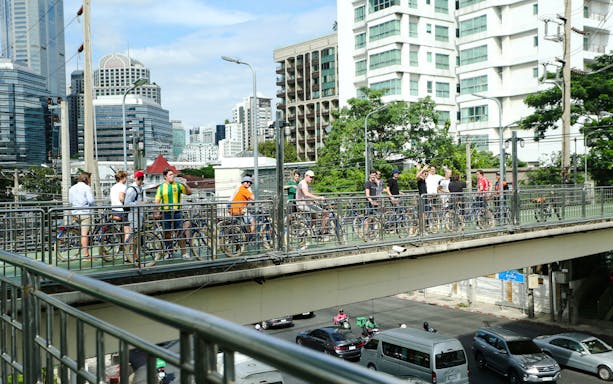 Cyclists crossing a footbridge in Bangkok city with skyscrapers in the background.