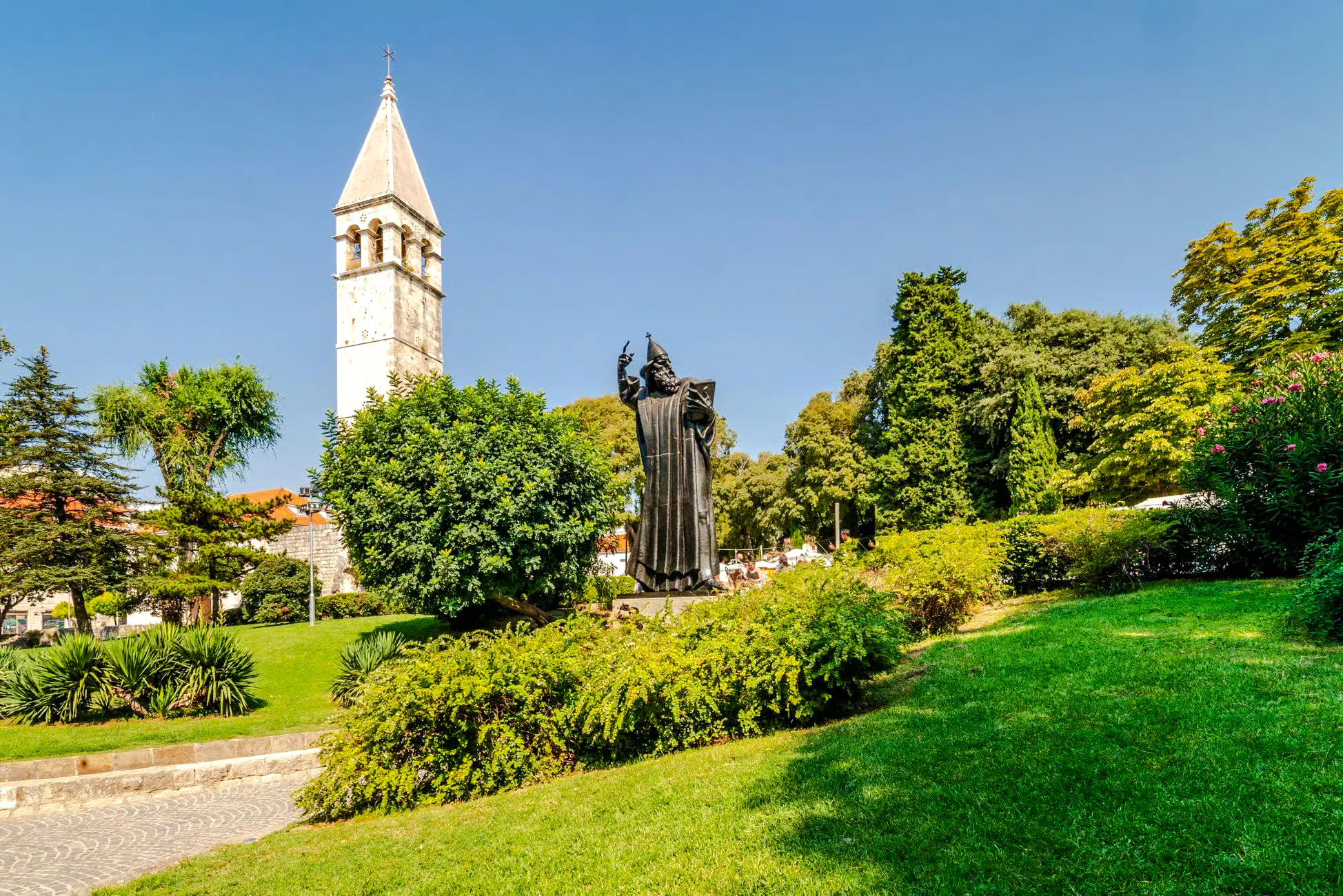 Statue of Grgur Ninski near bell tower in Split, Croatia, surrounded by greenery.