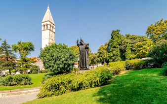 Statue of Grgur Ninski near bell tower in Split, Croatia, surrounded by greenery.