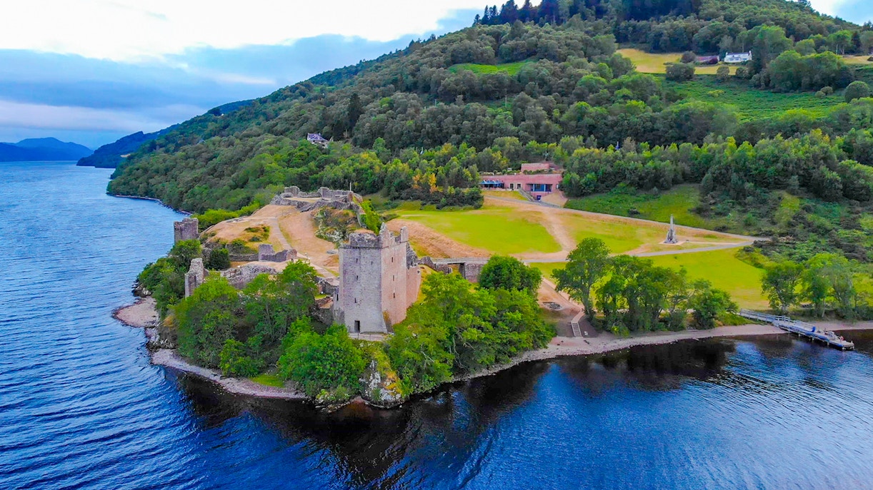 Aerial view of Urquhart Castle by Loch Ness in the evening, surrounded by lush greenery.