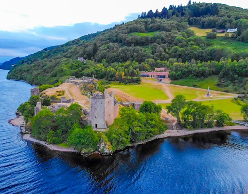 Aerial view of Urquhart Castle by Loch Ness in the evening, surrounded by lush greenery.
