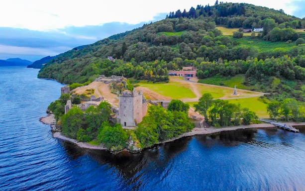 Aerial view of Urquhart Castle by Loch Ness in the evening, surrounded by lush greenery.