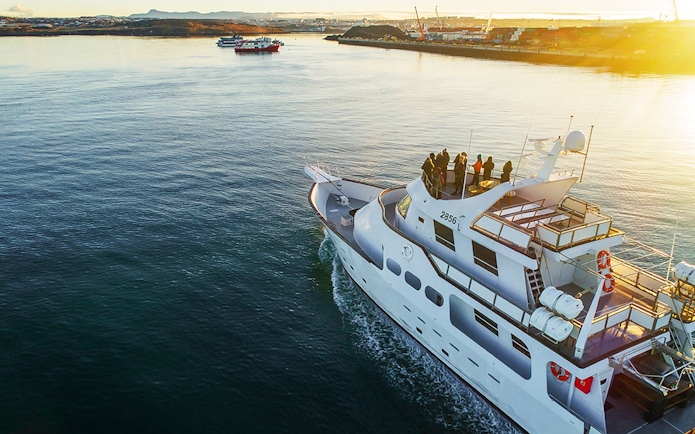 Luxury yacht cruising near a coastal city at sunset, passengers on deck enjoying the view.