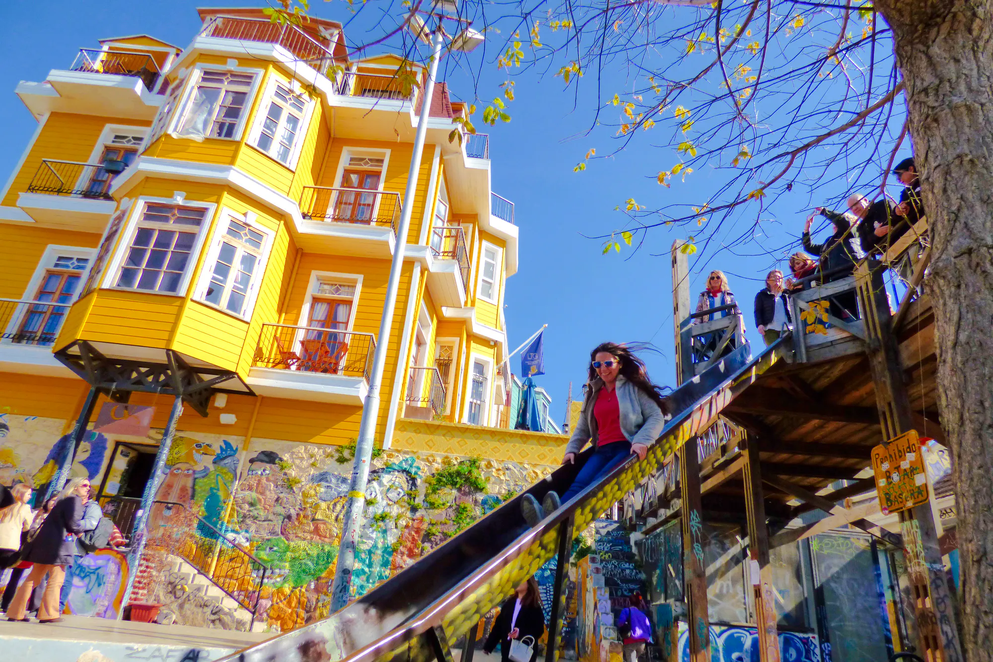 Woman sliding down a colorful slide in Valparaíso, Chile, near vibrant murals and a yellow building.