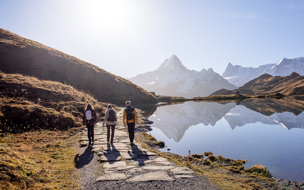 Hikers walking along a path by a lake with snow-capped mountains at Grindelwald First.