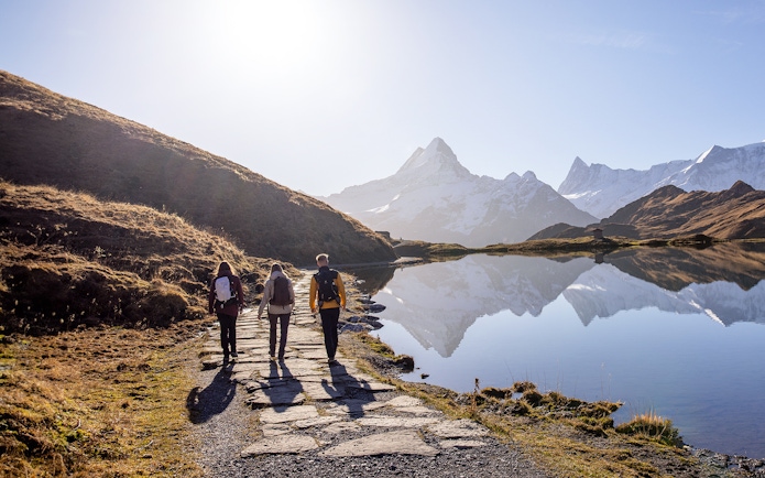 Hikers walking along a path by a lake with snow-capped mountains at Grindelwald First.