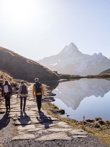 Hikers walking along a path by a lake with snow-capped mountains at Grindelwald First.