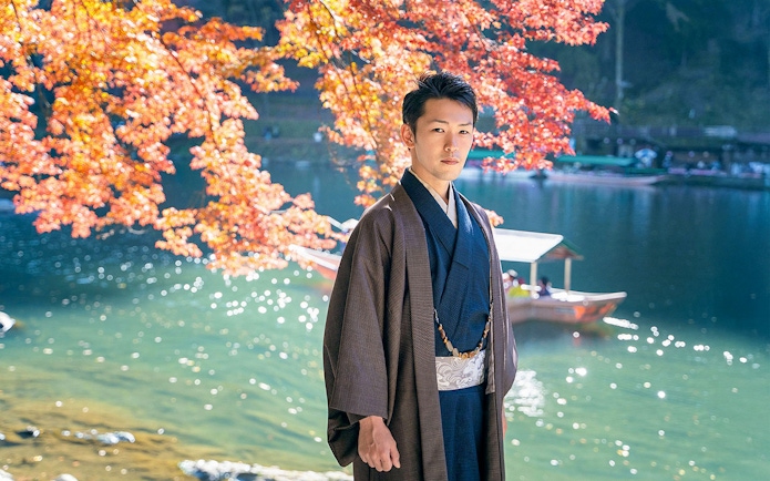 Man in kimono by river with autumn leaves in Kyoto Arashiyama.