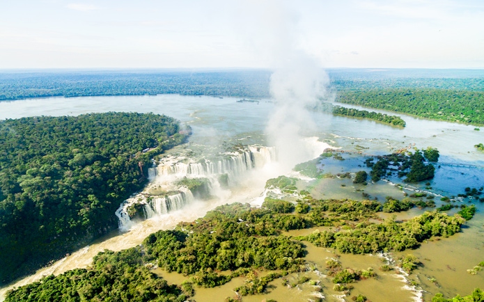 Aerial view of Iguazu Falls with surrounding lush forest and mist rising from the waterfalls.