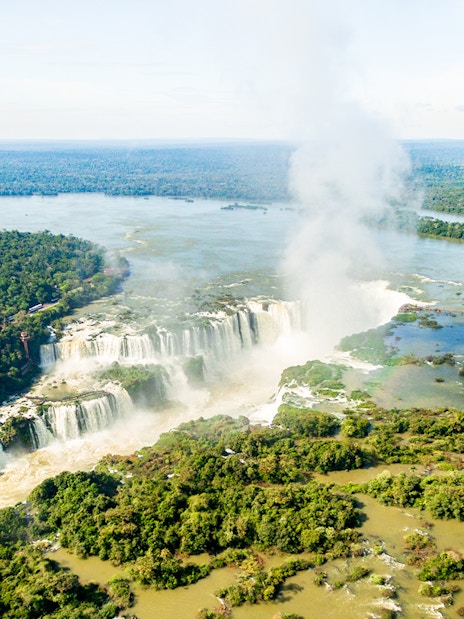 Aerial view of Iguazu Falls with surrounding lush forest and mist rising from the waterfalls.