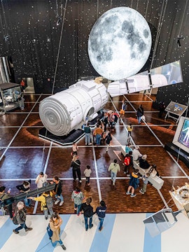 Visitors exploring space exhibit at Science & Industry Museum, Paris, featuring a large satellite model.