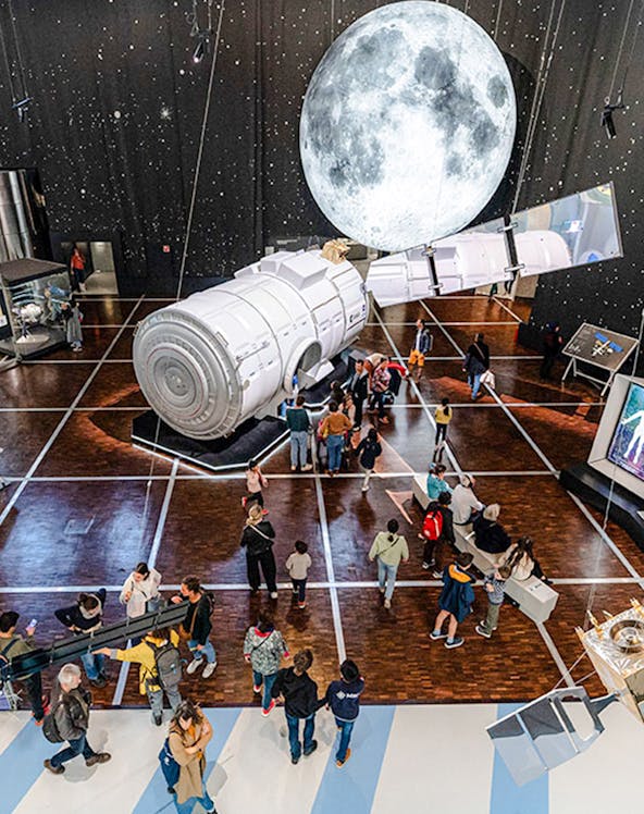 Visitors exploring space exhibit at Science & Industry Museum, Paris, featuring a large satellite model.