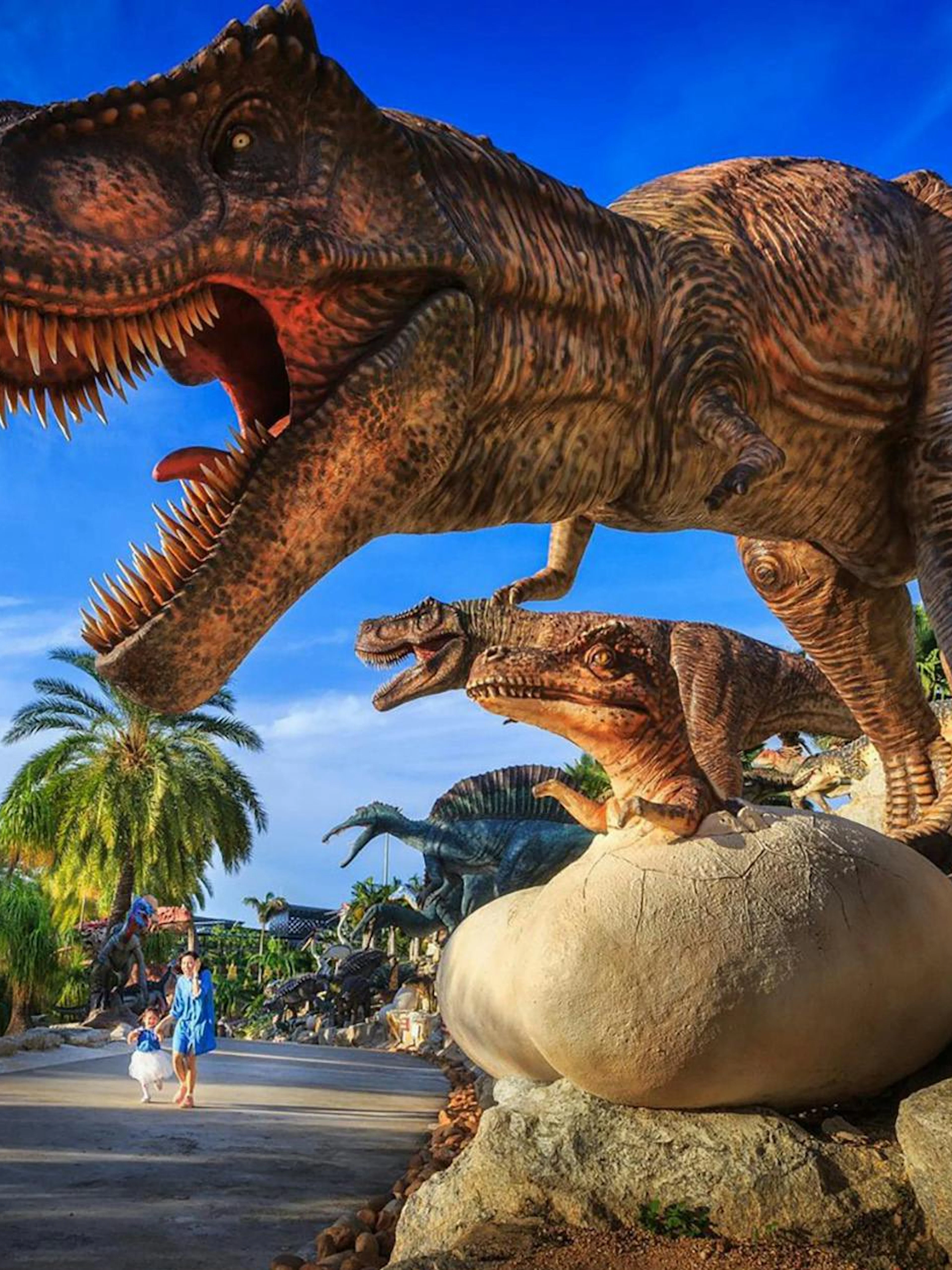 Dinosaurs at Nong Nooch Tropical Garden, Thailand, with palm trees and visitors.
