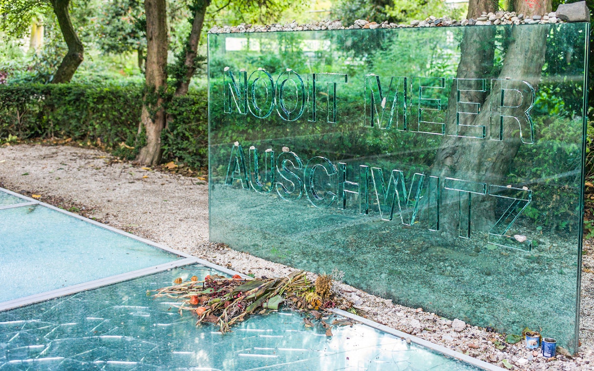 Auschwitz Memorial glass panel with stones and flowers in Amsterdam park.