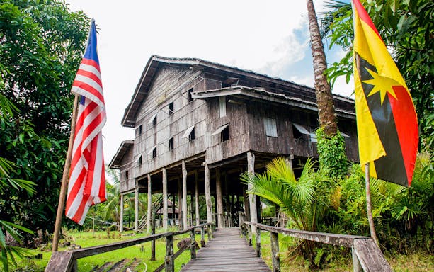 Traditional longhouse at Sarawak Cultural Village, Malaysia, with flags and lush greenery.