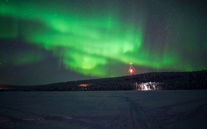 Northern lights over snowy landscape in Rovaniemi with distant forest and tower.