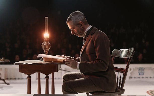 Man reading at a desk by lamplight during "An Enemy of the People" performance.