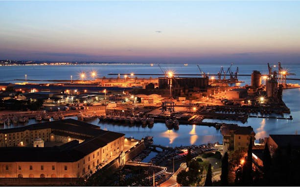 Ancona harbor at dusk with illuminated buildings and cranes by the waterfront.