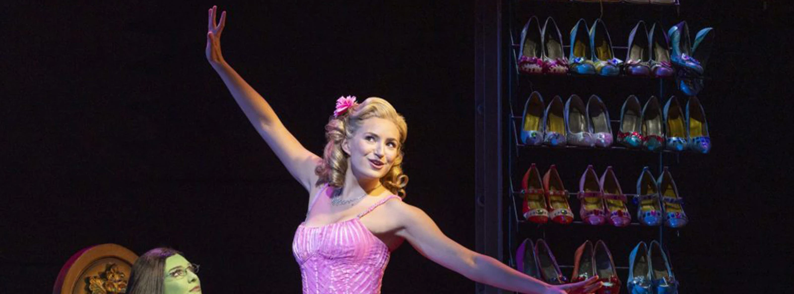Performers on stage in Wicked the Musical, with a shoe rack in the background.