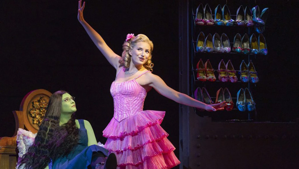 Performers on stage in Wicked the Musical, with a shoe rack in the background.