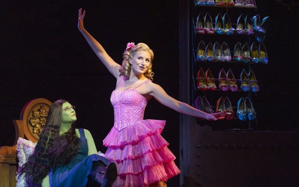 Performers on stage in Wicked the Musical, with a shoe rack in the background.