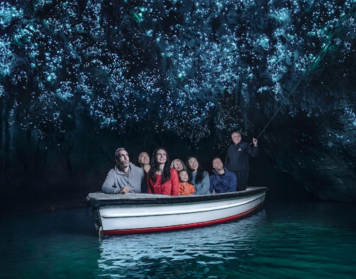 Visitors on a boat tour in Waitomo Glowworm Caves, New Zealand, admiring glowing cave ceiling.