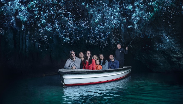 Visitors on a boat tour in Waitomo Glowworm Caves, New Zealand, admiring glowing cave ceiling.