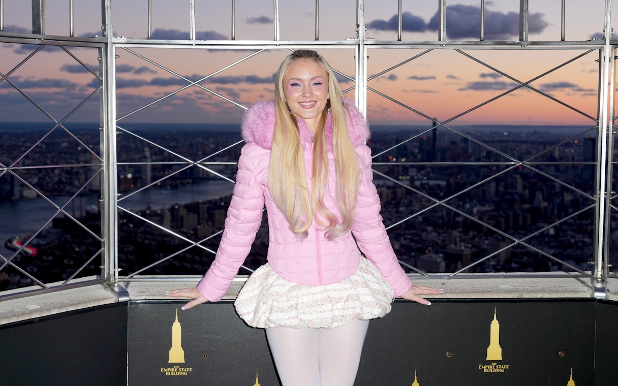Person in pink jacket at Empire State Building observatory, New York City skyline at sunset.