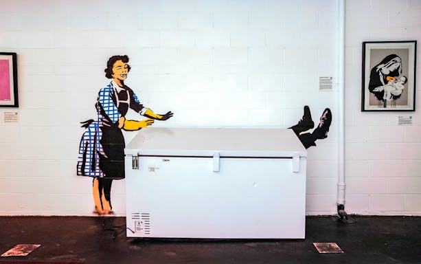 Banksy artwork of a woman pushing a freezer with legs sticking out, part of The Art of Banksy exhibit.