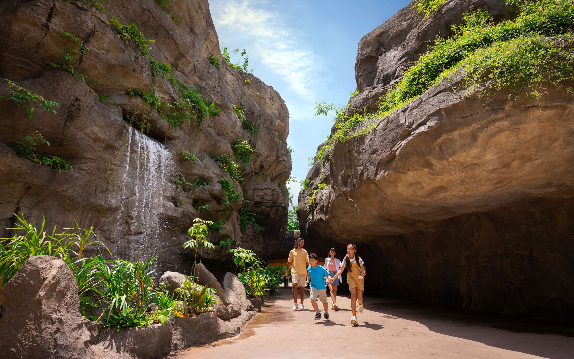 Family walking through Entrance Gorge at Rainforest Wild Adventure with waterfall and lush greenery.