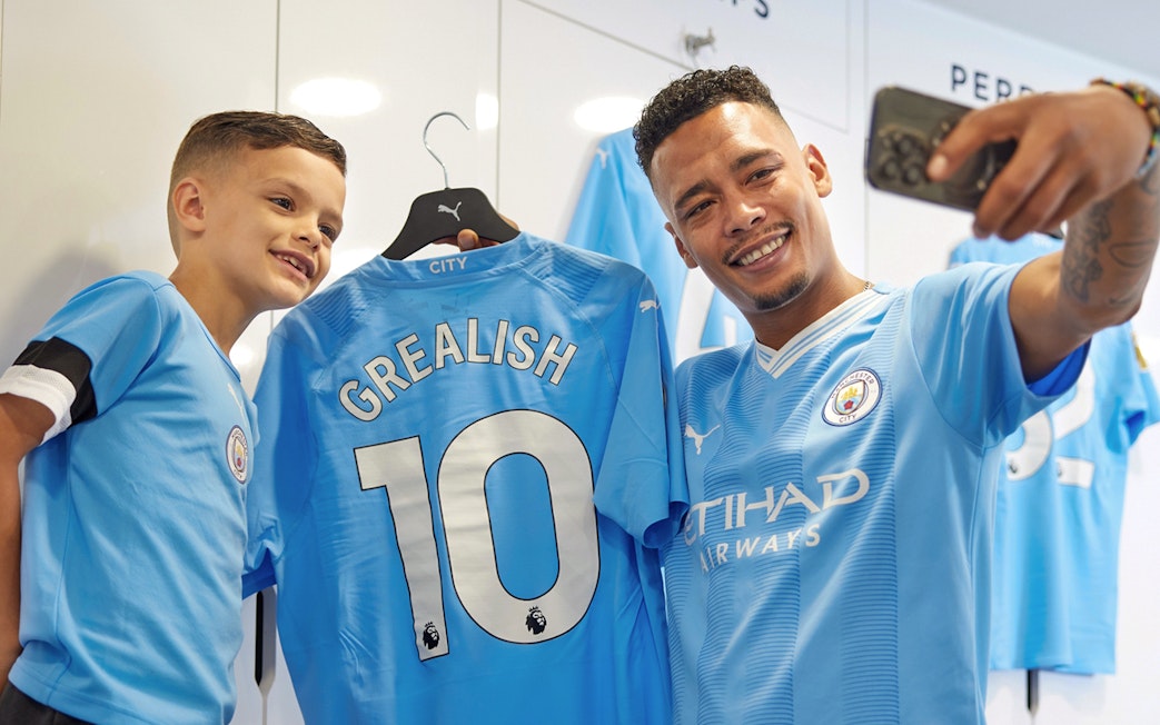 Man and child taking selfie with Manchester City jersey in stadium locker room.