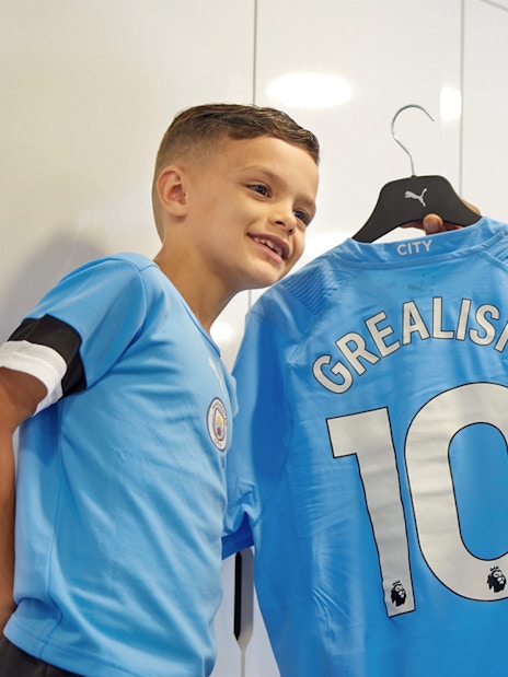 Man and child taking selfie with Manchester City jersey in stadium locker room.