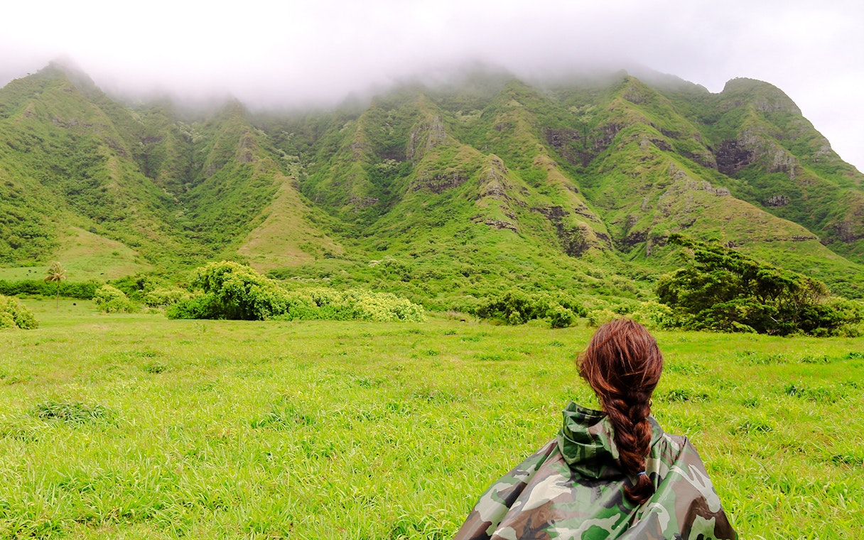 Person in camouflage jacket facing lush green mountains at Kualoa Ranch, Hawaii.