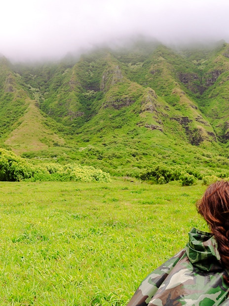 Person in camouflage jacket facing lush green mountains at Kualoa Ranch, Hawaii.