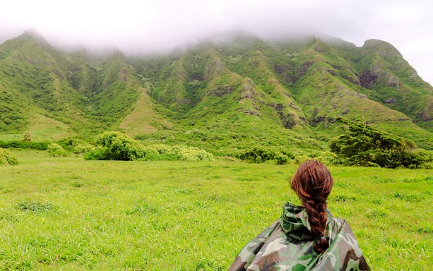 Person in camouflage jacket facing lush green mountains at Kualoa Ranch, Hawaii.