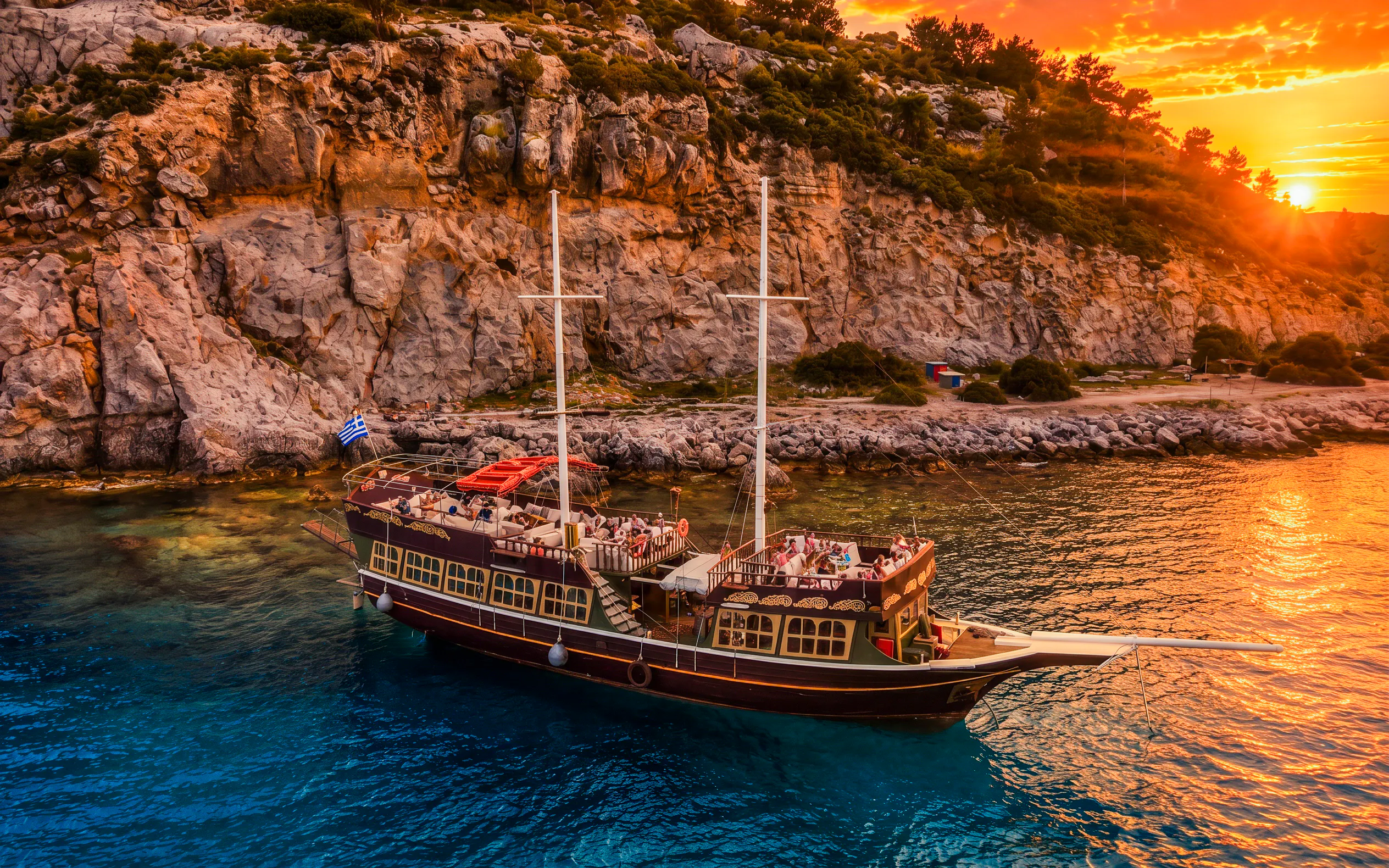 Sunset cruise ship near rocky coast of Rhodes with passengers enjoying drinks.