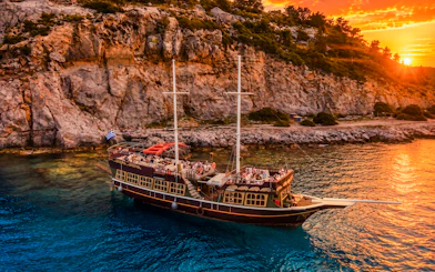 Sunset cruise ship near rocky coast of Rhodes with passengers enjoying drinks.