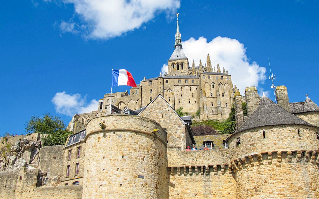 Mont Saint-Michel with French flag, stone walls, and abbey spire under blue sky.