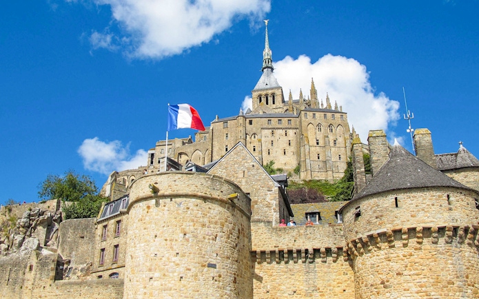 Mont Saint-Michel with French flag, stone walls, and abbey spire under blue sky.