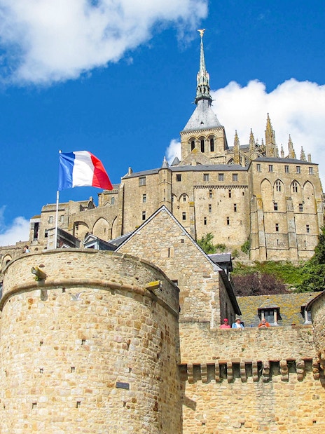 Mont Saint-Michel with French flag, stone walls, and abbey spire under blue sky.