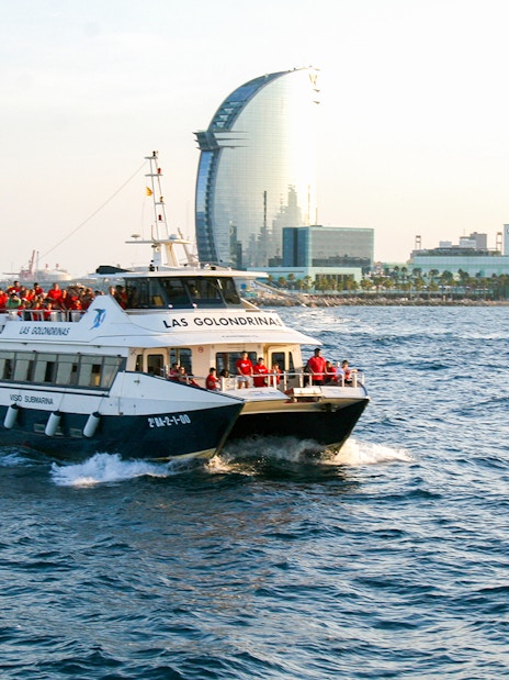 Las Golondrinas boat cruising near Barcelona waterfront with city skyline in background.