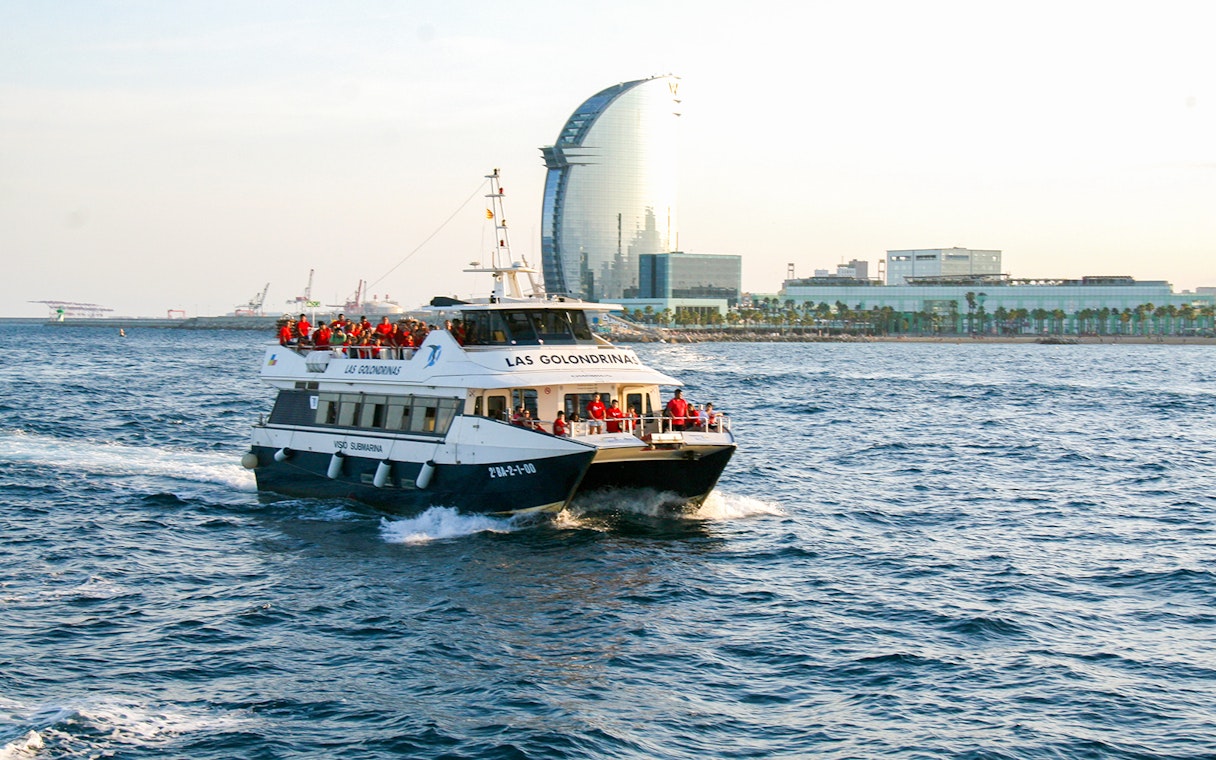 Las Golondrinas boat cruising near Barcelona waterfront with city skyline in background.