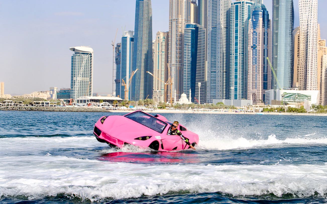 Man driving pink jet car on water with Dubai skyline in background.