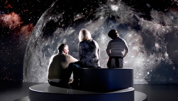 Visitors seated in front of a large moon projection at Illuminarium.
