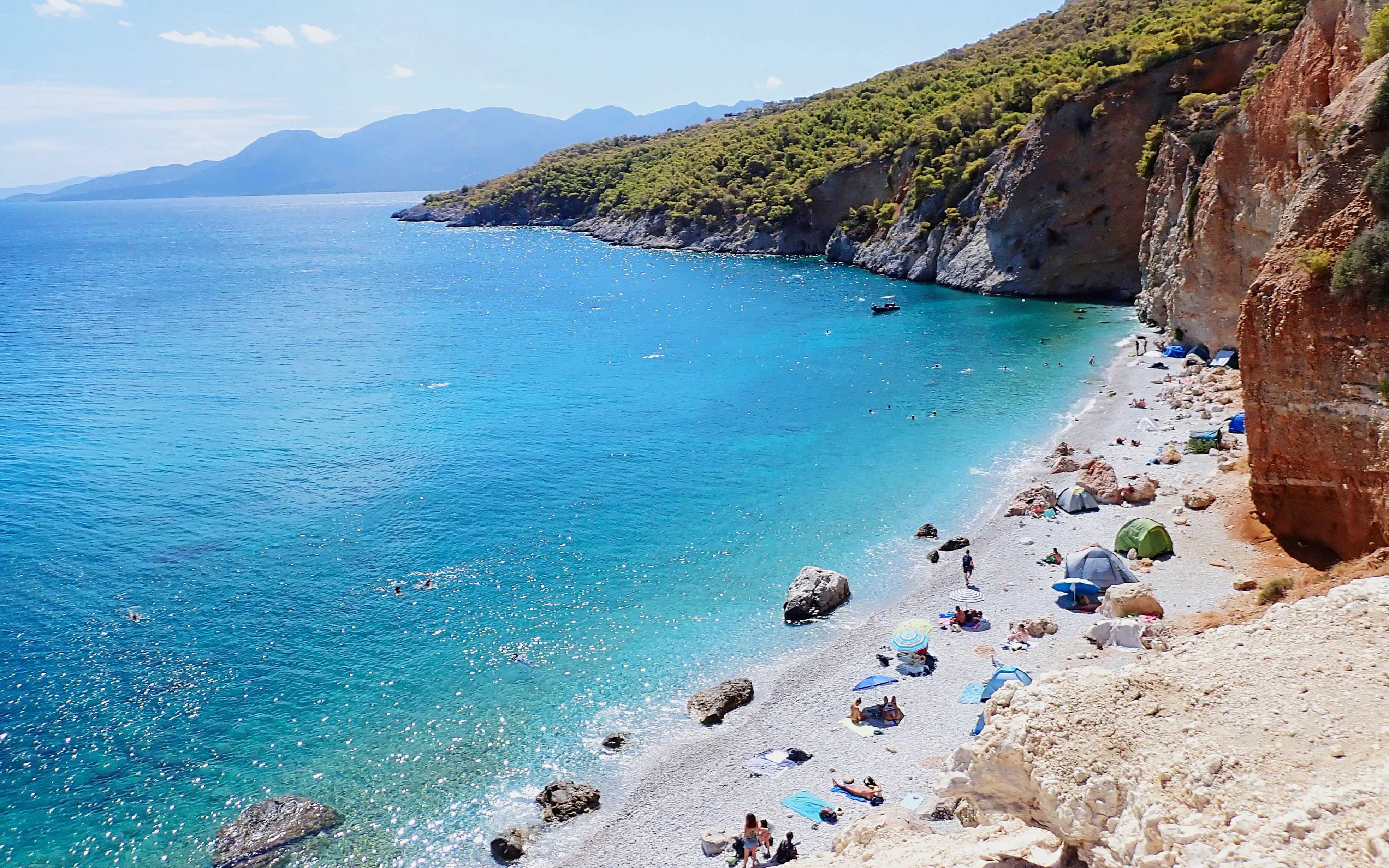 Chalikiada Beach on Agistri Island, Greece, with people sunbathing and swimming near rocky cliffs.