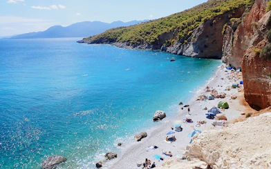 Chalikiada Beach on Agistri Island, Greece, with people sunbathing and swimming near rocky cliffs.
